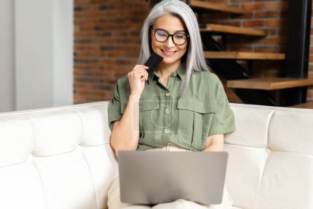 Photo for Pretty senior businesswoman in glasses sitting at the sofa using laptop and credit card for online payment from home, typing number to enter internet banking account, ordering food - Royalty Free Image