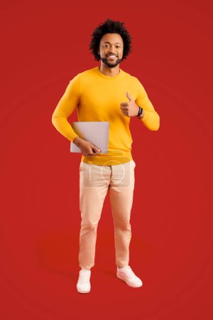 Téléchargez les photos : Homme souriant tenant un ordinateur portable dans les mains et souriant, bel homme indépendant debout isolé sur fond rouge - en image libre de droit