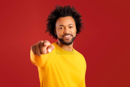 Foto de Alegre hombre afroamericano señala con el dedo índice a la cámara aislada en el fondo rojo, guapo tipo multirracial eligiéndote, indicando con una sonrisa - Imagen libre de derechos