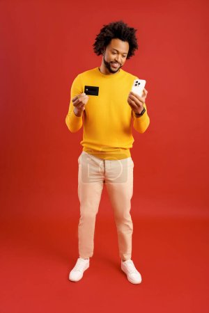 Téléchargez les photos : E-banking, transfert d'argent en ligne. homme afro-américain joyeux tenant carte de crédit et smartphone dans ses mains isolé sur rouge, noir en utilisant l'application mobile pour le paiement en ligne, achats - en image libre de droit
