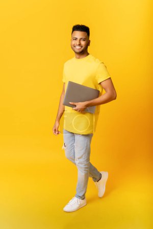 Photo for Cheerful Indian man carrying laptop computer isolated on yellow. Happy arab male student with laptop walking, freelancer using portative gadget, computer. Vertical photo - Royalty Free Image