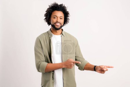 Foto de Pleasant looking curly man pointing finger aside at copy space, showing area for advertisement. Indoor studio shot isolated on white background - Imagen libre de derechos