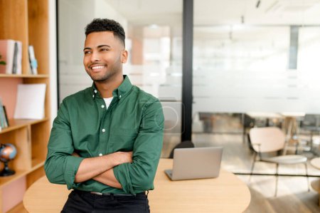 Thriving Indian male employee stands confidently with his arms folded, showcasing a positive and cheerful smile. Freelancer working in smart casual shirt in shared workspace