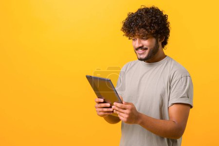 Foto de Sonriente guapo indio joven rizado hombre leyendo libro electrónico, utilizando tableta digital para navegar por la web o estudiar a la distancia, foto de estudio aislado en amarillo, espacio de copia - Imagen libre de derechos