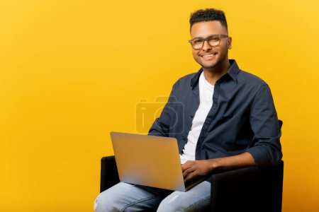 Foto de Bonito empresario indio sonriente que usa un portátil sentado en el sillón aislado en amarillo, retrato de un estudiante masculino feliz y determinado mirando la cámara con la computadora en su regazo, hombre árabe escribiendo - Imagen libre de derechos