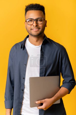 Foto de Retrato del exitoso hombre indio en gafas de pie y portando portátil, mirando a la cámara aislada en amarillo, alegre empresario masculino, freelancer o estudiante sonriendo - Imagen libre de derechos