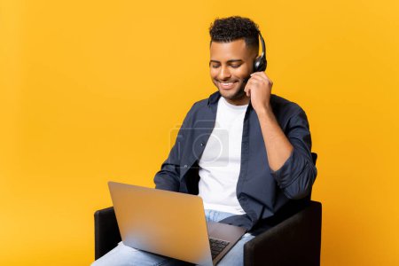 Photo for In touch. Smiling young indian man in headset using laptop, salesman talking online, positive man working in the customer service department, isolated on yellow, taking call - Royalty Free Image