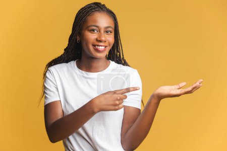 Photo for Positive woman pointing at copy space on her palm, empty place for idea presentation, product advertising. Indoor studio shot isolated on yellow background - Royalty Free Image