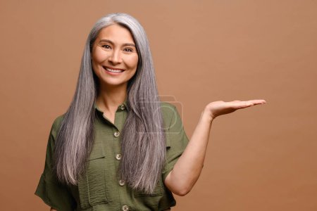 Photo for Serene middle-aged 40s asian woman holding palm open for new product or sale presentation standing isolated on brown, charming mature female advertising nevelty looking at camera - Royalty Free Image
