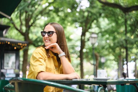 Foto de Encantadora morena con gafas de sol y camisa amarilla pasando tiempo en la terraza de la cafetería. Mujer joven serena y despreocupada disfrutando del café de la mañana al aire libre - Imagen libre de derechos