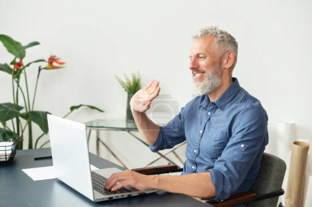 Photo for Cheerful mature grey-haired man using laptop for video call sitting at the desk in the office, senior male looks at the webcam, smiling and waving. Virtual meeting concept. Side view - Royalty Free Image