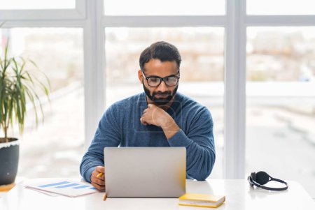 Photo for Concentrated Indian guy is typing on the laptop, answering emails, chatting with coworkers. Clever and focused ethnic businessman in casual and stylish glasses wear in office space - Royalty Free Image