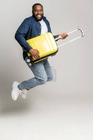 Foto de Alegre chico afroamericano saltando con una maleta amarilla, juega en una bolsa como un guitarrista, tonteando, un hombre negro con el equipaje mirando hacia adelante un viaje, aislado en gris, longitud completa, vertical - Imagen libre de derechos