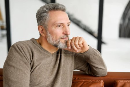 Photo for Portrait of serene and carefree handsome middle-aged grey-haired man sitting on the couch and looks aside lost in thoughts and dreams. Peaceful well-looking mature male resting at home alone - Royalty Free Image