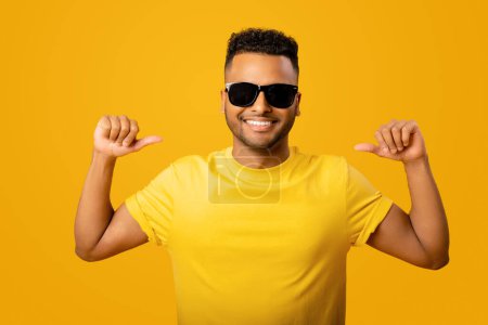 Téléchargez les photos : Souriant jeune homme indien joyeux en lunettes de soleil et t-shirt debout isolé sur fond jaune pointant vers lui-même, choix d'un homme ethnique confiant, publicité - en image libre de droit