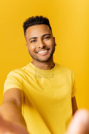 Téléchargez les photos : Charmant homme indien prenant selfie, portant un t-shirt décontracté souriant regardant joyeusement la caméra debout isolé sur fond jaune, pov homme arabe faire un appel vidéo, enregistrement vidéo - en image libre de droit