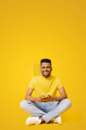 Photo for Smiling indian man using smartphone sitting on the floor in relaxed pose, isolated on yellow backdrop, carefree serene male student chatting, spends time in social networks, scrolling news feed - Royalty Free Image