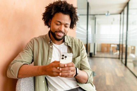 Photo for Smiling african american business man with smartphone in hand looking screen and sitting indoor, male office employee, entrepreneur using smartphone, chatting and texting online - Royalty Free Image