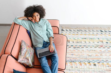 Téléchargez les photos : Jeune femme d'affaires noire ou pigiste dans des vêtements décontractés confortables assis sur le canapé et travaillant sur le projet, tapant un e-mail sur l'ordinateur portable, écrivant un article, regarde la caméra, souriant - en image libre de droit