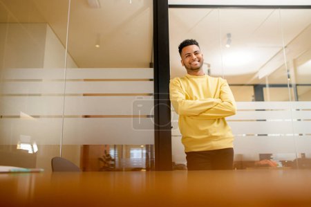 Téléchargez les photos : Succès confiant jeune brunet indien adulte entrepreneur homme d'affaires professionnel en vêtements décontractés, debout avec les bras croisés pliés au bureau moderne et regardant la caméra - en image libre de droit