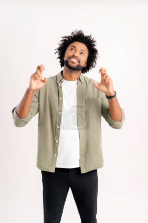 Photo for Wish you good luck. Handsome 30s curly guy in casual shirt holding fingers crossed isolated on white, charismatic african-american man hoping for miracle, strong wishing something, praying - Royalty Free Image