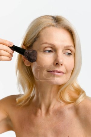 Téléchargez les photos : Portrait vertical de charmante blonde des années 40 50 femme d'âge moyen appliquant le maquillage avec brosse isolée sur blanc - en image libre de droit