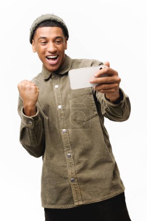 Photo for Excited young man playing video game on the smartphone isolated on white background, hispanic gambling guy spends leisure time with video games, celebrating victory, creaming yes - Royalty Free Image