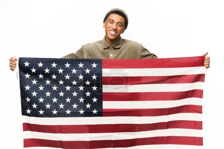 Foto de Alegre chico afroamericano sosteniendo en las manos la bandera americana, mirando a la cámara y sonriendo aislado sobre fondo blanco, concepto del Día de la Bandera - Imagen libre de derechos