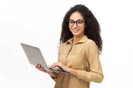Foto de Atractiva mujer afroamericana joven en gafas y camisa casual inteligente de pie y usando computadora portátil aislada sobre fondo blanco, empleado multirracial usando aplicación informática - Imagen libre de derechos
