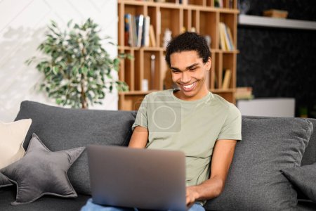 Photo for Cheerful multiracial man in casual wear is sitting on the comfortable sofa and using trendy laptop for video call, speaking with interlocutor online. Virtual meeting, video call concept - Royalty Free Image