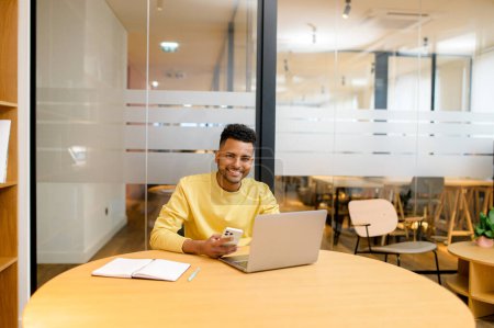 Photo for Portrait of positive indian male student holding smartphhone and sitting at desk in front of laptop and working on a project in office, happy man looking at the camera with cheerful smile, using phone - Royalty Free Image