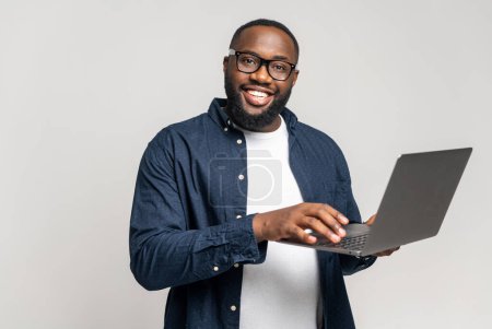 Téléchargez les photos : Sourire homme africain en utilisant un ordinateur portable, debout à fond gris, vérifier les finances de la maison, homme heureux regardant la facture, regardant la caméra, connexion avec la banque, vérifier la facture en utilisant l'application en ligne - en image libre de droit