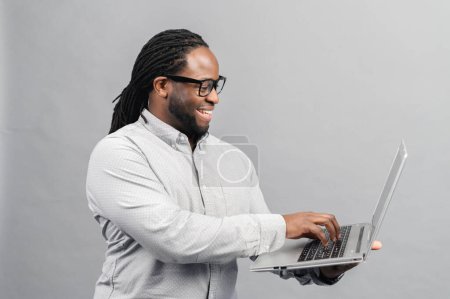 Téléchargez les photos : Jeune homme afro-américain debout isolé sur le mur gris tenant ordinateur portable et le message de dactylographie, bavarder avec un ami sur les médias sociaux en ligne, en utilisant sortir ensemble app, profiter de la nouvelle application - en image libre de droit