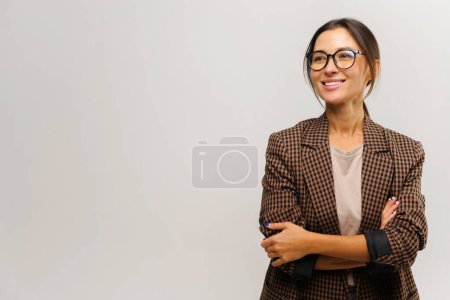 Téléchargez les photos : Portrait de mignonne femme d'affaires positive portant un costume formel debout avec les bras croisés et regardant loin avec un sourire charmant. Studio intérieur tourné isolé sur fond blanc - en image libre de droit