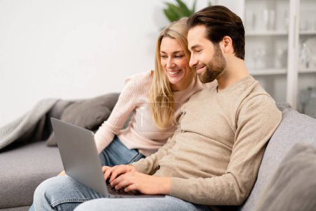 Overjoyed couple in love spending leisure time online with laptop at home. Blonde woman and man looking at the laptop screen while sitting on the sofa and watching comedy movies Poster 653486524
