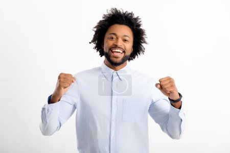 Foto de Emocionado joven afroamericano en camisa blanca formal posando aislado sobre fondo blanco, levantando puños en gesto ganador, espacio de copia - Imagen libre de derechos
