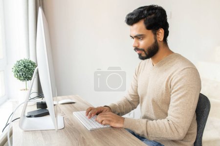 Photo for Optimistic ethnic man in casual wear using computer sitting at the table. Young indian male student watching webinars, educational courses, learning on the distance, typing emails - Royalty Free Image