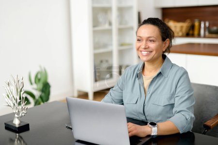 Photo for Confident middle-aged woman work with a laptop at the modern home office. Female employee in smart casual wear looks at the camera and smiles, typing messages, answering email, front view - Royalty Free Image
