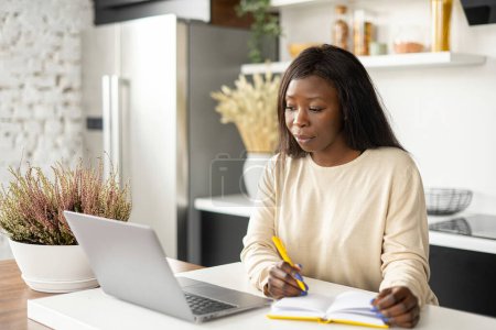 Téléchargez les photos : Une belle femme noire en tenue décontractée intelligente utilise un ordinateur portable dans un bureau à domicile moderne, une employée écrit dans un planificateur, une étudiante étudie à distance, regarde un webinaire - en image libre de droit