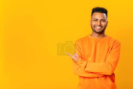 Photo for Advertising concept. Cheerful indian guy point finger aside, presenting novelty, paying your attention at empty copy space, brazilian in orange pullover isolated on yellow - Royalty Free Image