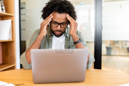 Téléchargez les photos : Inquiète collègue afro-américain employé masculin regardant l'écran d'ordinateur portable assis sur le lieu de travail, cher homme d'affaires concerné a un problème avec le projet, manque de nouvelles idées - en image libre de droit