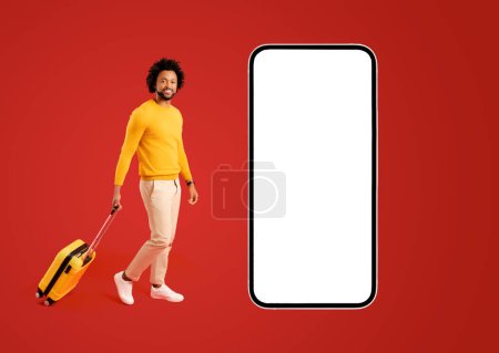 Photo for Traveler man with a yellow suitcase luggage walking near gigant smartphone with empty screen. Smiling carefree African-American guy isolated on red recommends app for travel and trips - Royalty Free Image