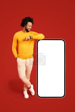 Photo for Best deal, promotion. Smiling positive african-american man leaning on huge smartphone with empty blank screen, looking at it, freelancer guy recommending new app or website, mockup - Royalty Free Image