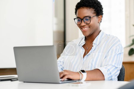 Foto de Amistosa y positiva empleada afroamericana o estudiante usando laptop, mujer de negocios mirando la pantalla, escribiendo, respondiendo a correos electrónicos sentados en la oficina contemporánea - Imagen libre de derechos