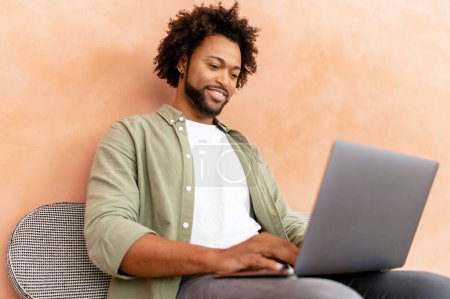 Téléchargez les photos : Jeune homme afro-américain positif assis dans le bureau de style loft et utilisant un ordinateur portable. Employé masculin joyeux tapant sur le clavier, regarde l'écran satisfait - en image libre de droit