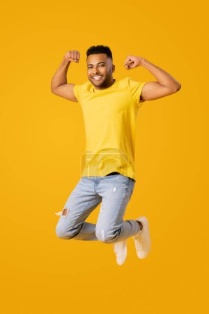Téléchargez les photos : Portrait complet de bonhomme indien athlétique heureux sautant haut et montrant biceps, jeune homme en tenue décontractée isolé sur fond de couleur jaune levant les poings en signe de victoire - en image libre de droit