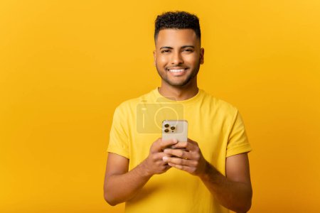 Foto de Retrato de un joven indio alegre enviando un mensaje en un teléfono móvil sobre un fondo amarillo. Hombre árabe feliz usando teléfono inteligente, disfrutando de chatear en línea, mensajería en las redes sociales - Imagen libre de derechos