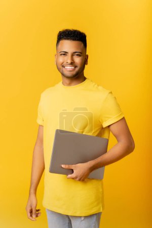 Téléchargez les photos : Un jeune indépendant indien souriant se tient dans un studio isolé sur un ordinateur portable jaune et branché. gai arabe guy dans casual t-shirt regarde caméra, tenant ordinateur portable fermé, étudiant - en image libre de droit