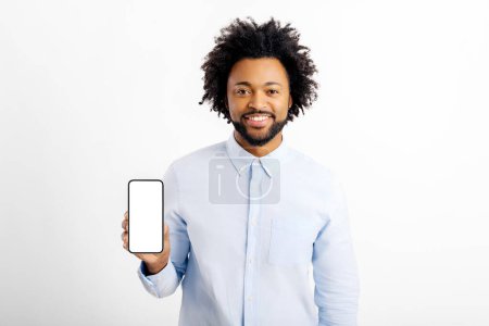 Foto de Amistoso y sonriente tipo multirracial que muestra el teléfono inteligente con la pantalla vacía en blanco, la presentación de la nueva aplicación móvil, empleado de oficina masculino en la publicidad de camisa formal blanca - Imagen libre de derechos