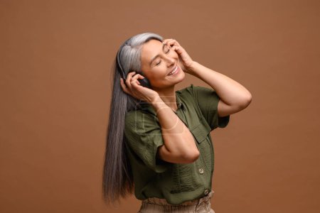 Téléchargez les photos : Serene femme asiatique d'âge moyen dans les écouteurs écoutant de la musique et dansant isolé sur fond brun, détendu souriant dame des années 50 aux cheveux gris appréciant les pistes préférées - en image libre de droit